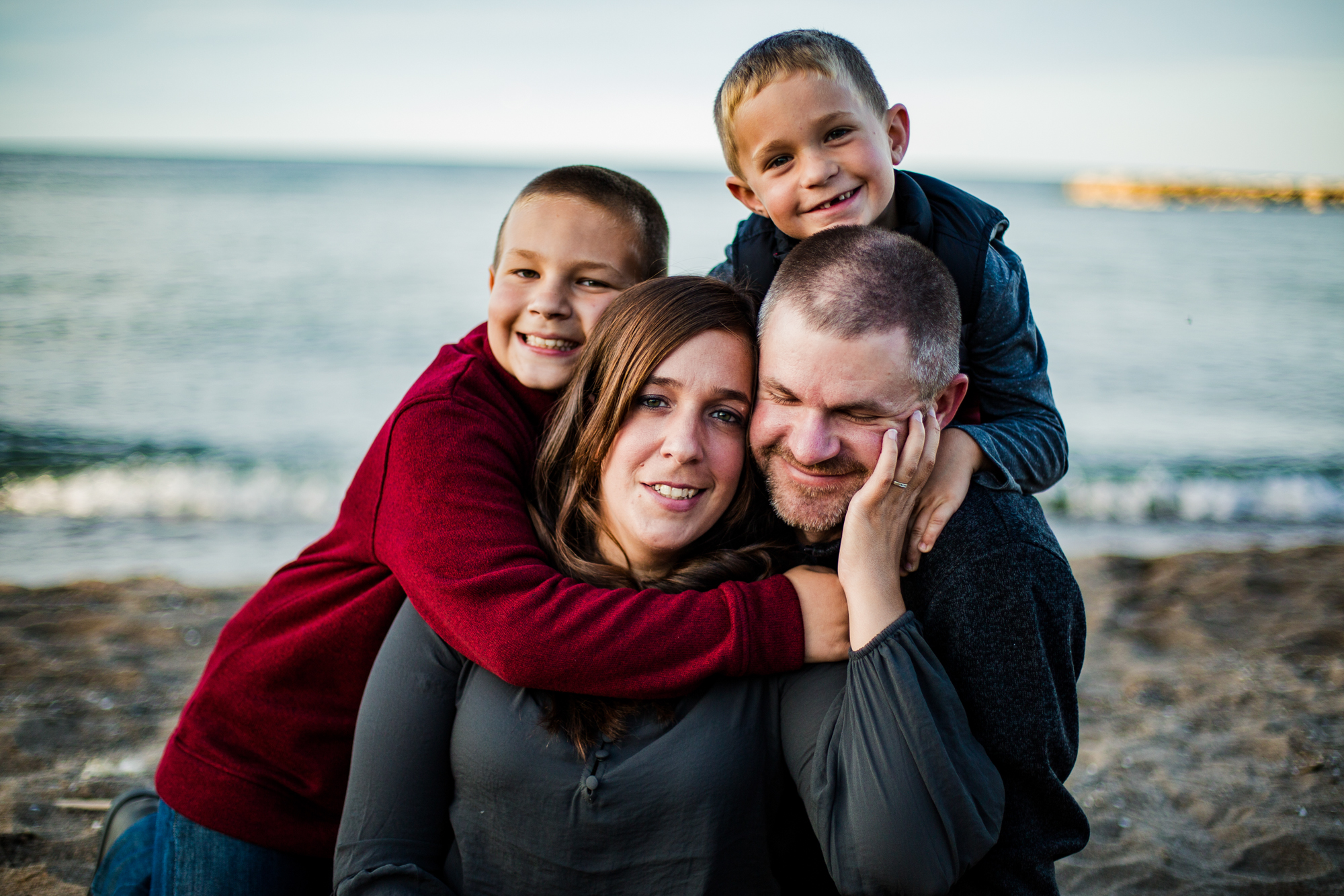 Wife cradles husband's face as their two sons lean on them on the beach at Presque Isle State Park