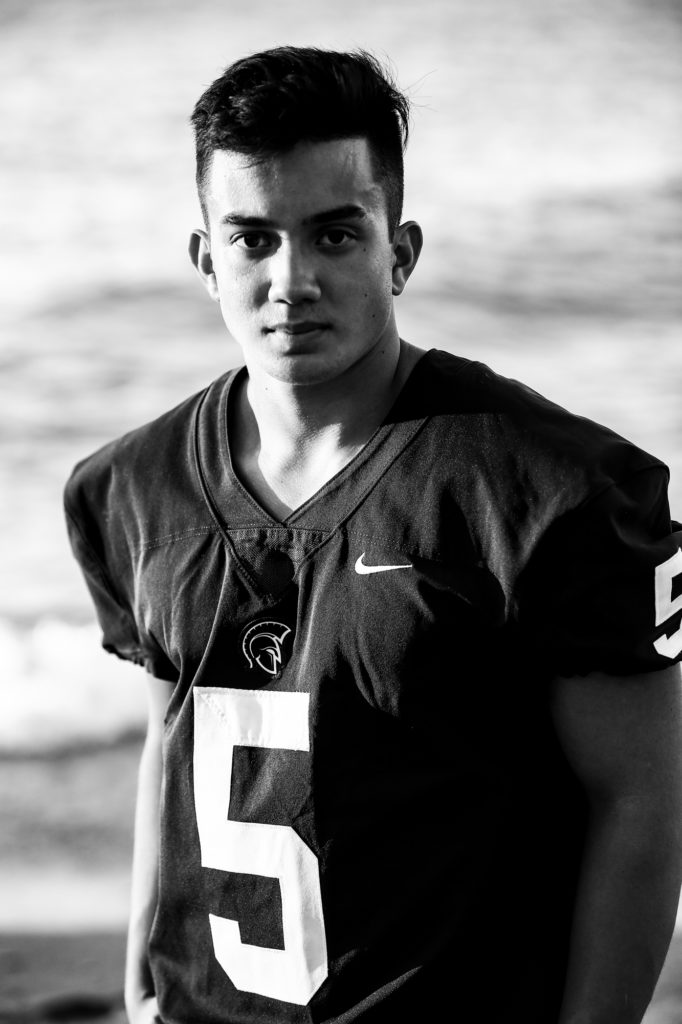 young man wearing football jersey stands next to Lake Erie for Presque Isle senior portraits