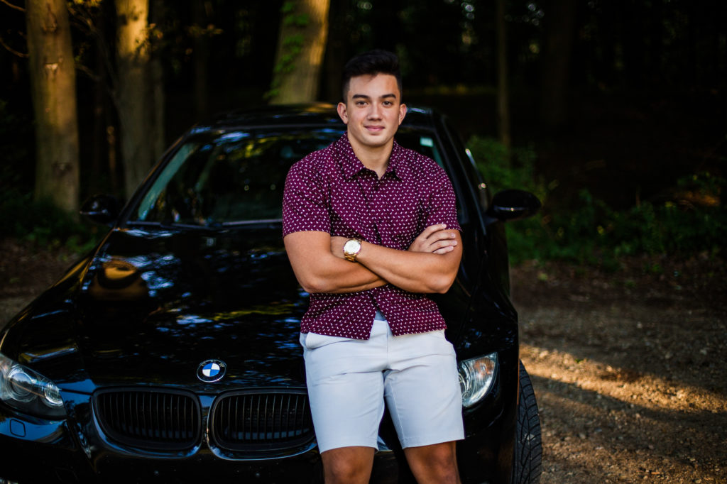 High school senior smiles while leaning against his car at Presque Isle State Park in Erie PA