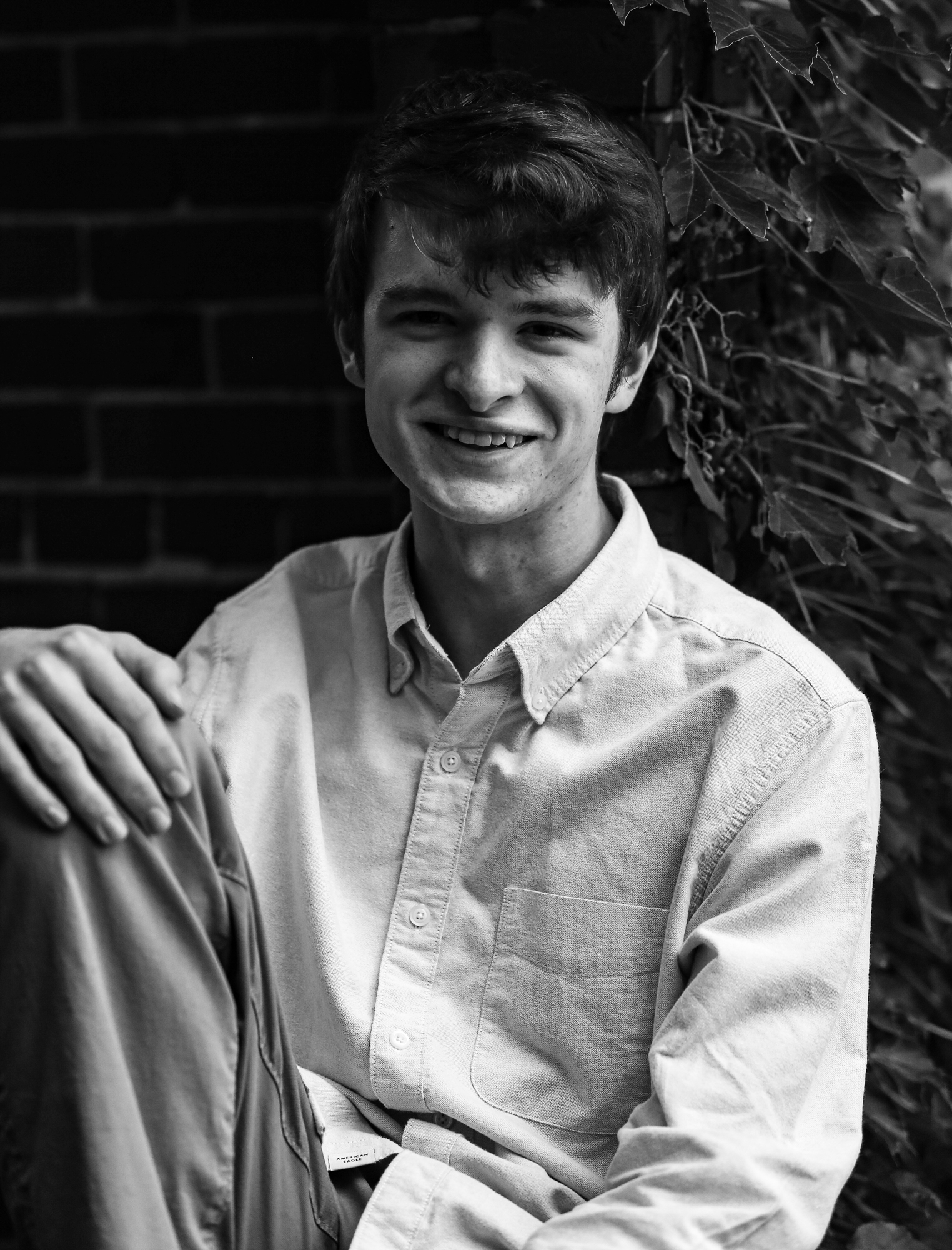 Young man smiling while sitting next to ivy covered brick wall at Modern Tooll Square during senior photos in Downtown Erie