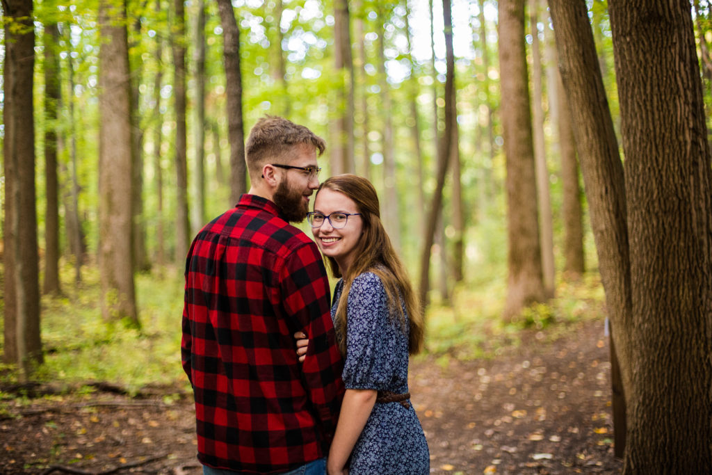 Woman looks back at camera as man kisses the top of her head for summer enagagement portraits at Asbury Woods