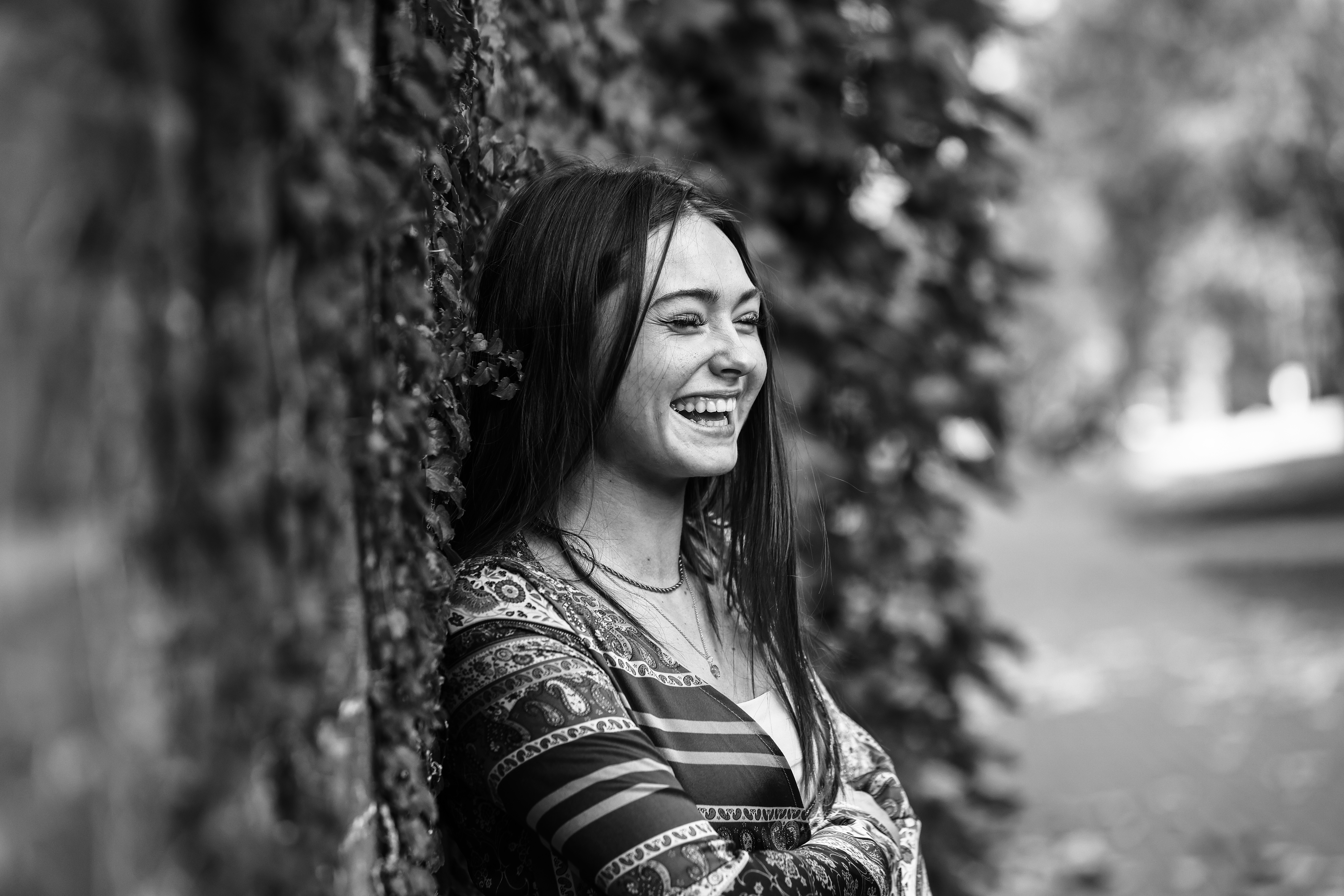 A young woman outside, laughing, while leaning against a brick wall covered in ivy.