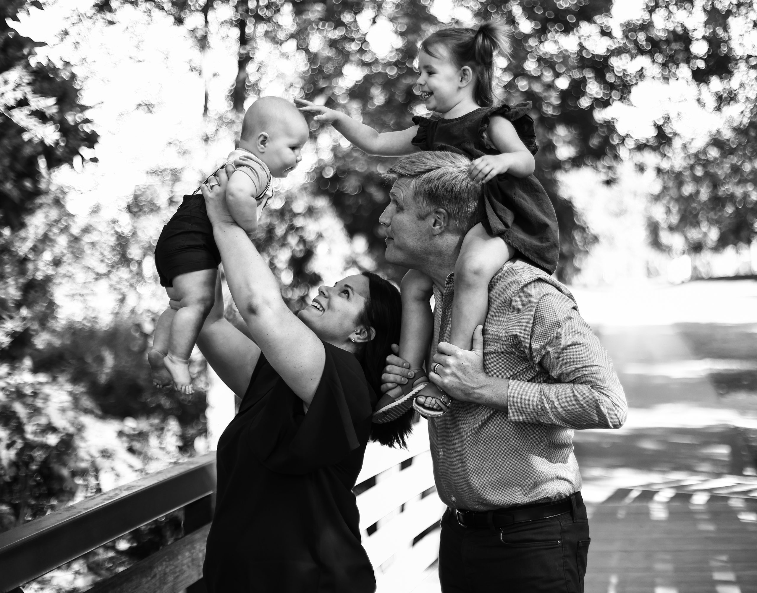 A woman and man, outdoors on a bridge, with two young children. The woman is holding the younger child above her and the man is holding the other child on his shoulders. How to look great in photos.