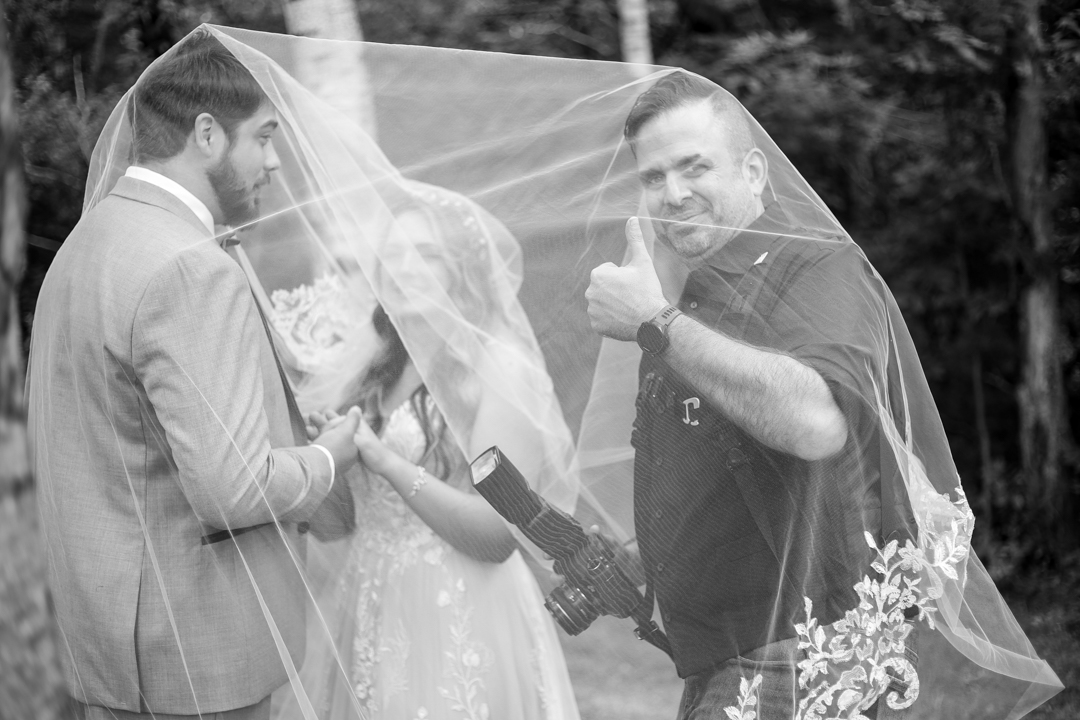Photography of a bride and groom under the veil with the photographer with his camera and giving the thumbs up.
