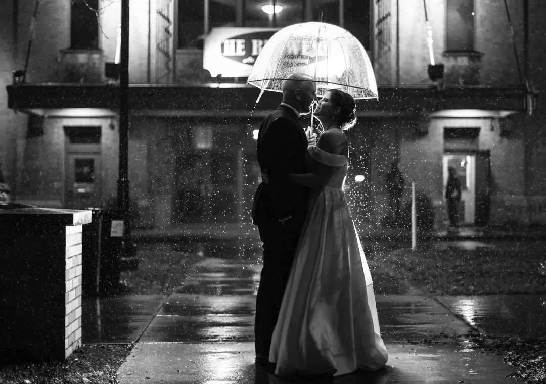 Photography of a bride and groom, outside in the rain, under a clear umbrella.